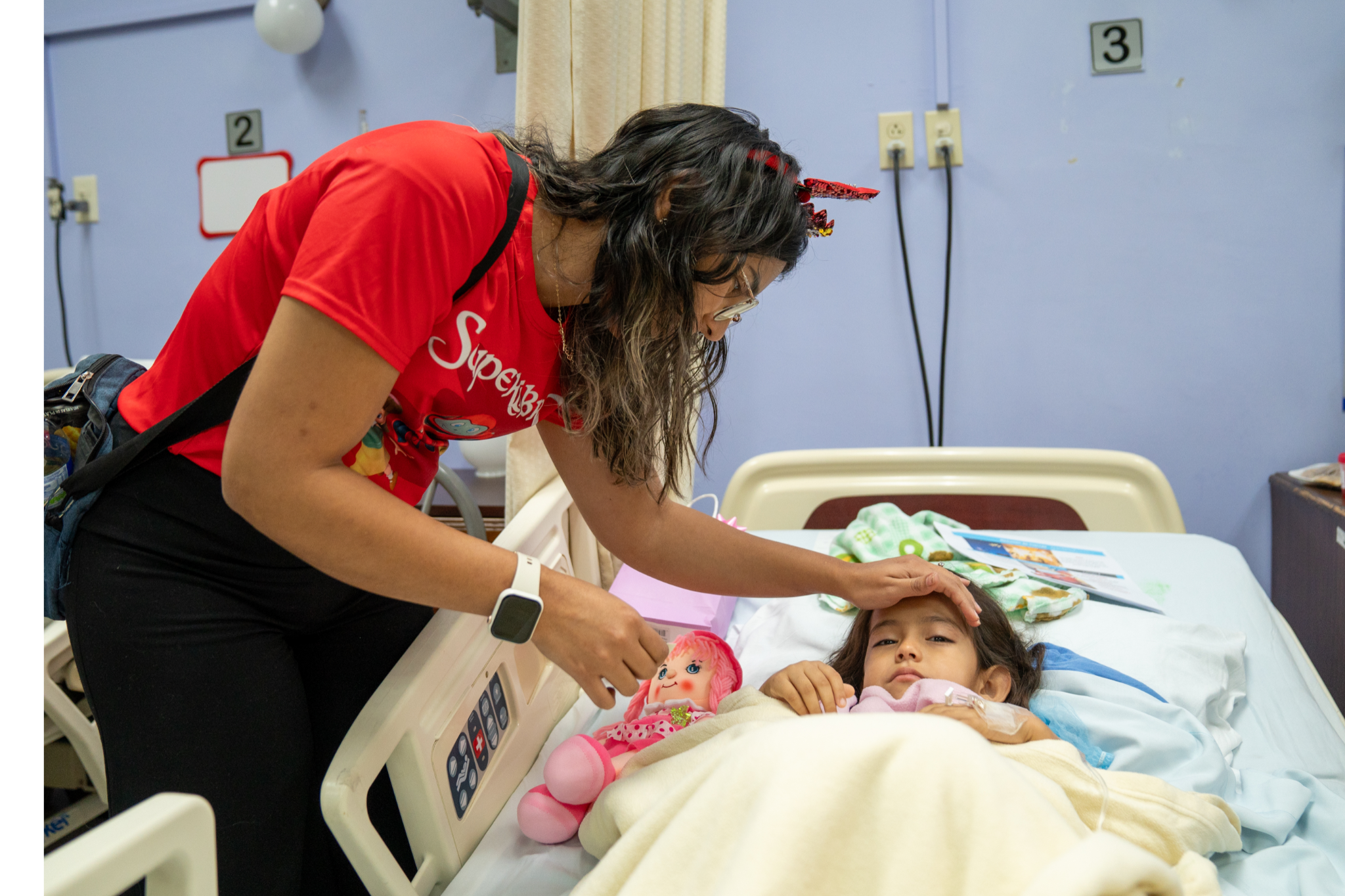 A young girl lays in a hospital bed while a Superbook team member comforts her.