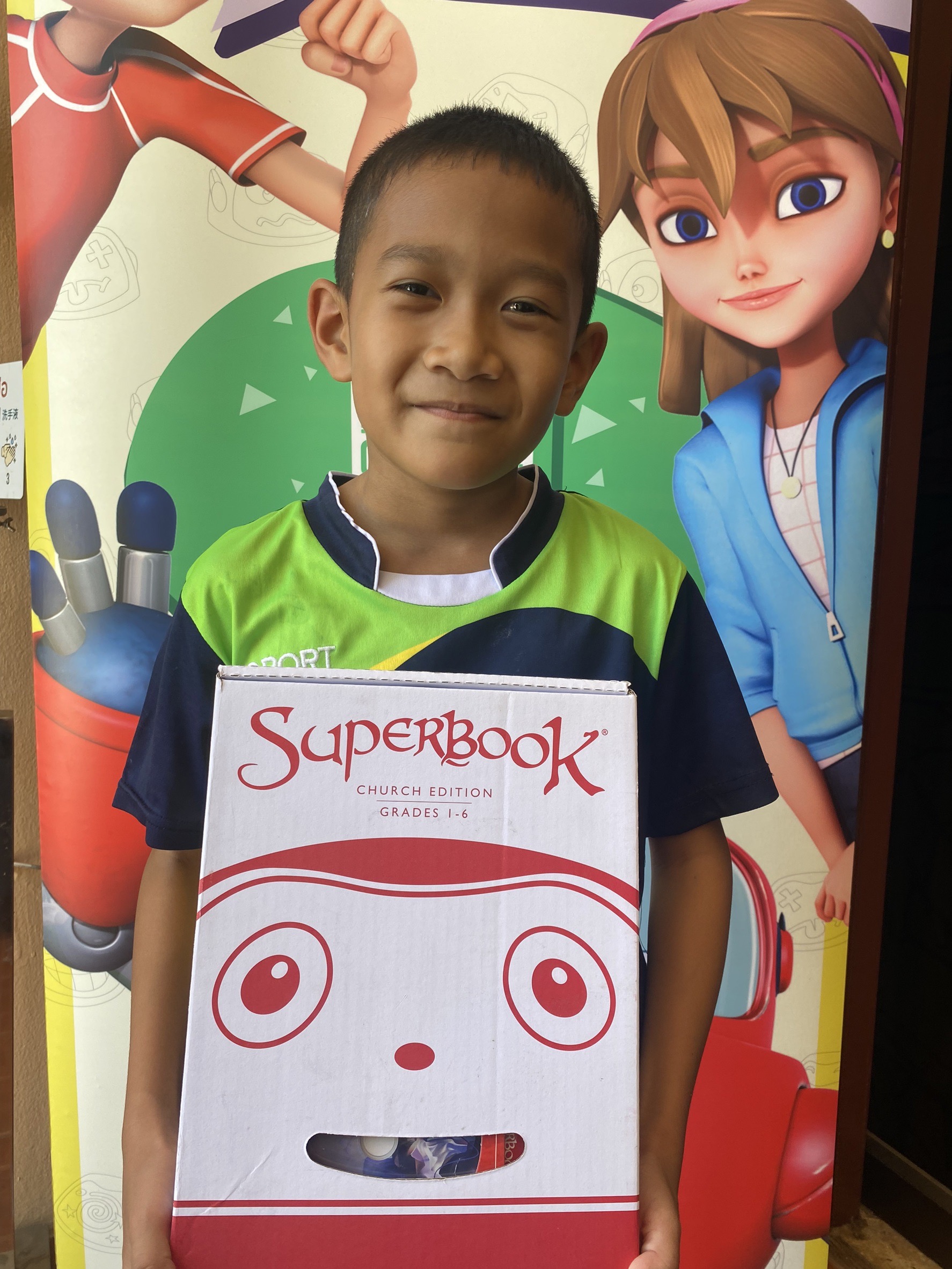 Ekkasit smiles at the camera, holding a Superbook Academy box