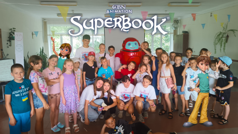 A group of children in Ukraine gather around Gizmo. Joy and Chris also stand with the children. The CBN Animation and Superbook logos are stacked in the top center of the photo.